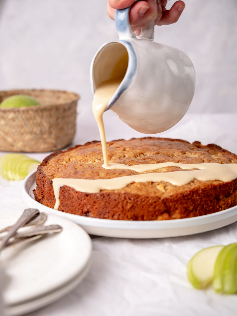 Vanilla Sauce being drizzled over a moist and delicious buttermilk apple cake with sliced apples on the side.