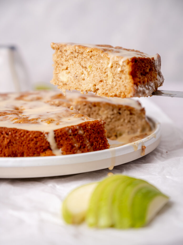 The perfect slice of cozy buttermilk apple cake being lifted on a cake server with vanilla drizzle.