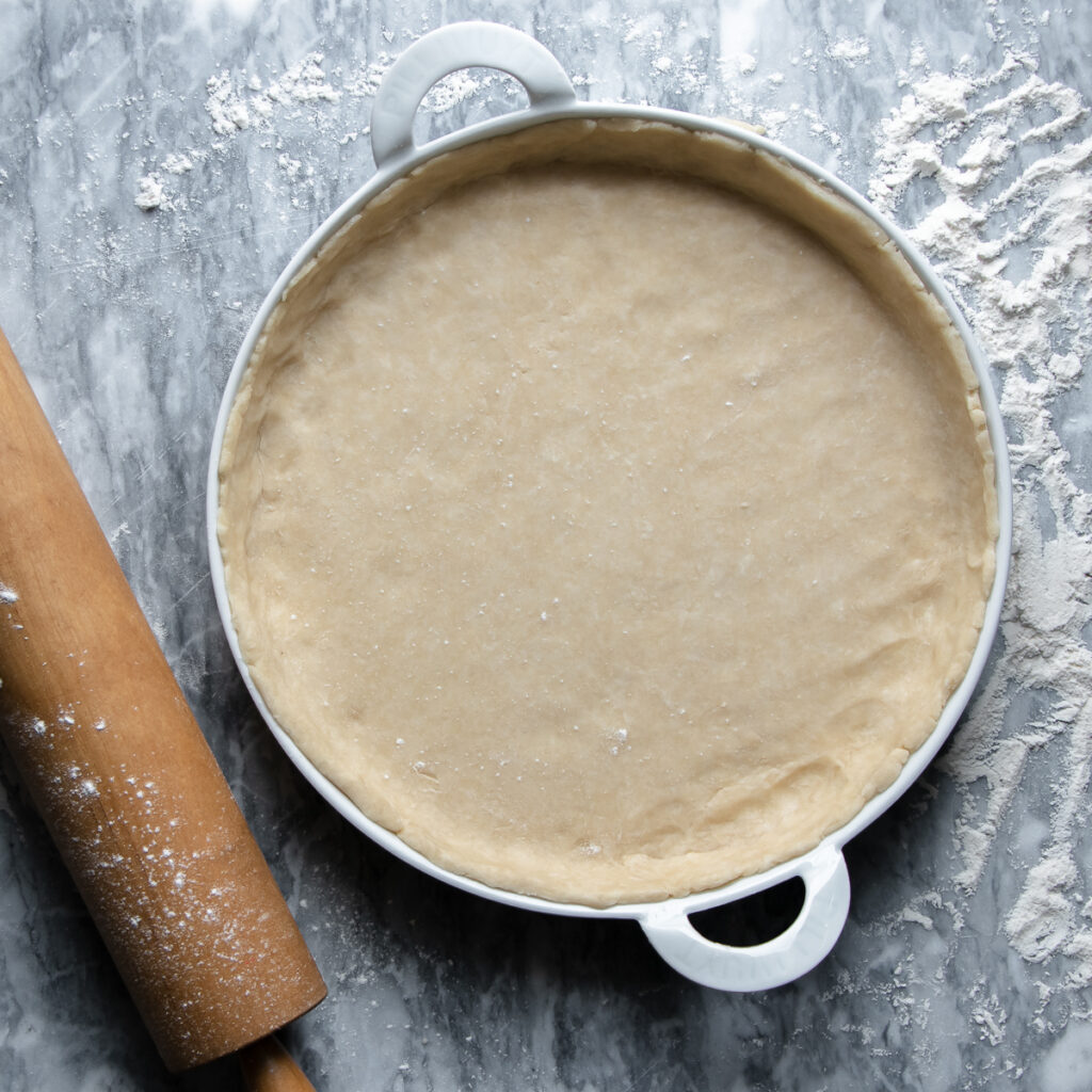 Unbaked quiche crust in a toulouse french porcelain tart pan made by Pillivuyt and a rolling pin next to it.