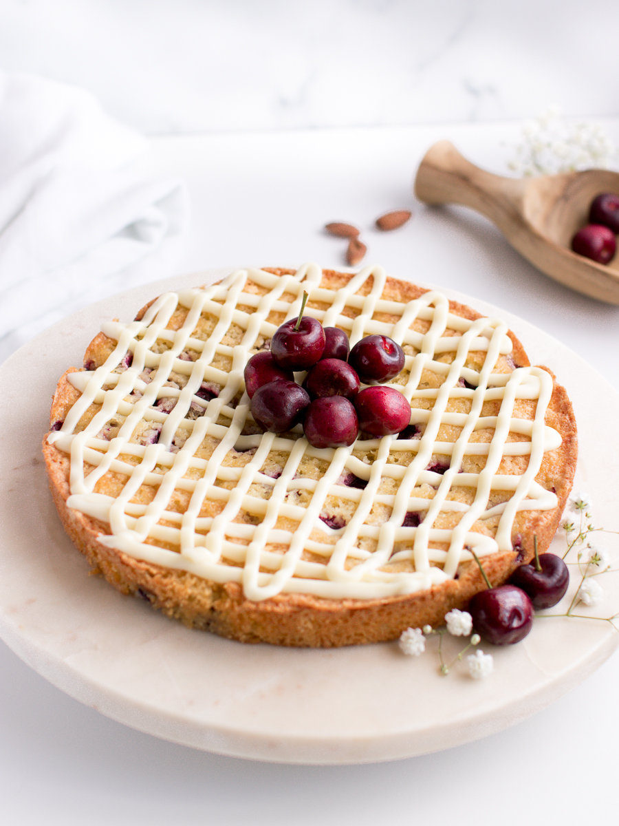 Simple recipe for almond cake, shown with cherries on the side and lattice-designed white chocolate drizzle on top.