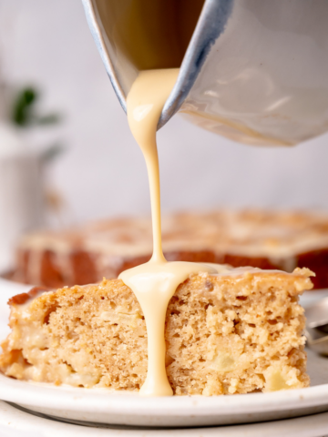 Creamy vanilla sauce slowly being poured over a slice of buttermilk apple cake.