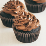 Three chocolate cupcakes with a rich swirl of chocolate frosting on top, sprinkled with chocolate shavings, displayed on a neutral background.