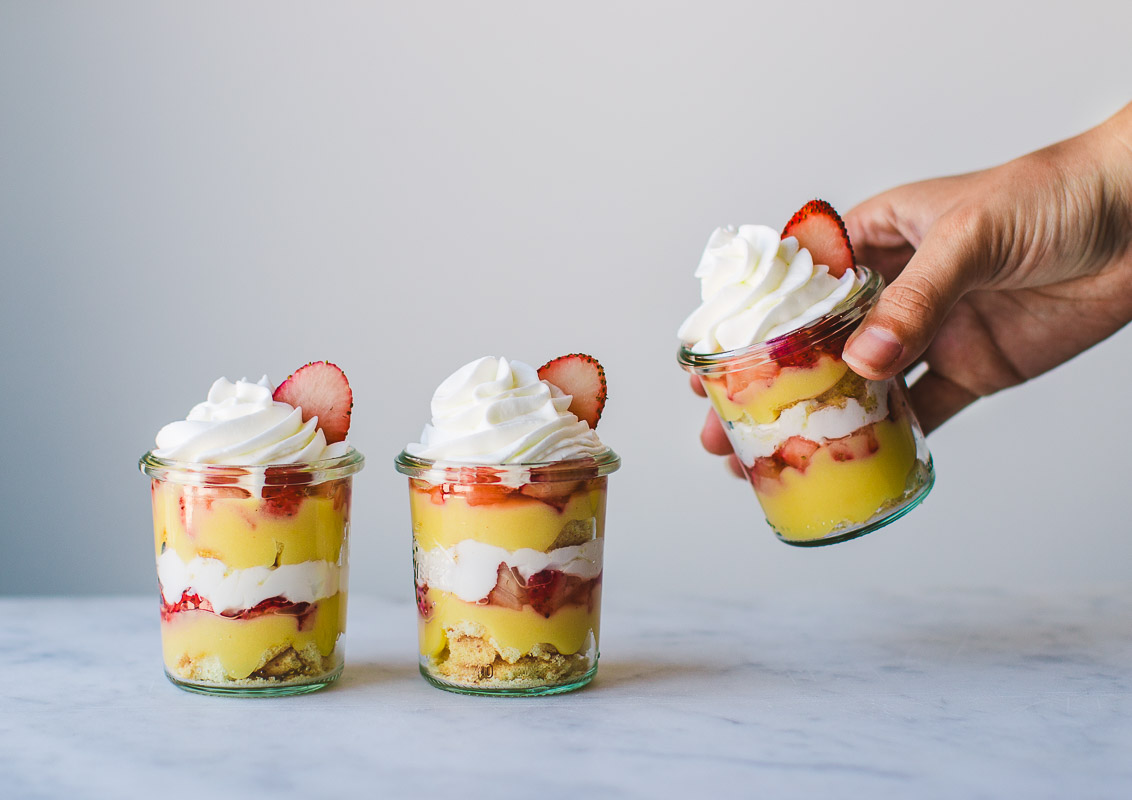 A hand grabbing 1 of 3 jars of Lemon curd Strawberry Parfaits with swirls of whipped cream and fresh strawberry garnish.