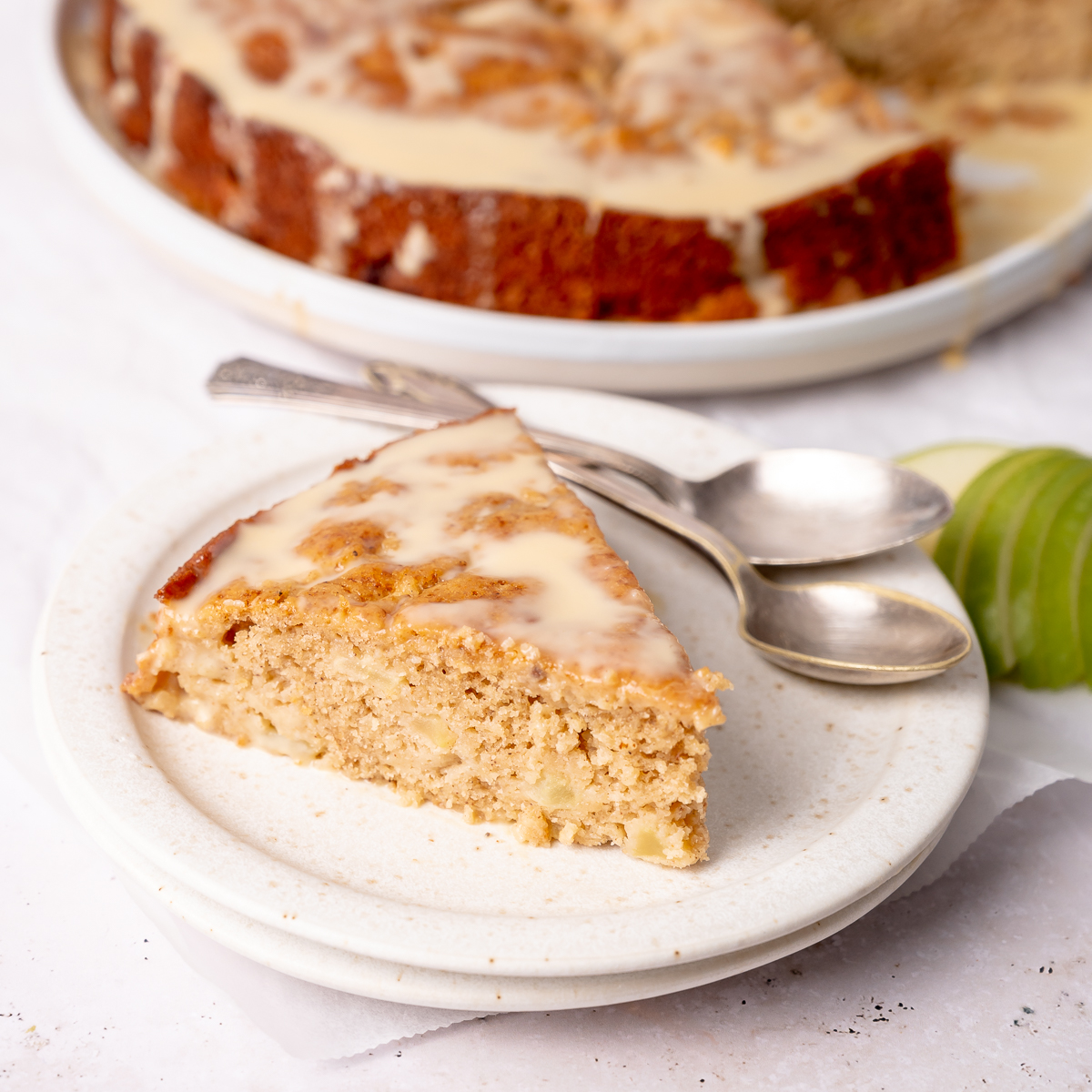 Two spoons on a plate with a fresh slice of apple cake and vanilla sauce drizzle with a sliced green apple on the side.