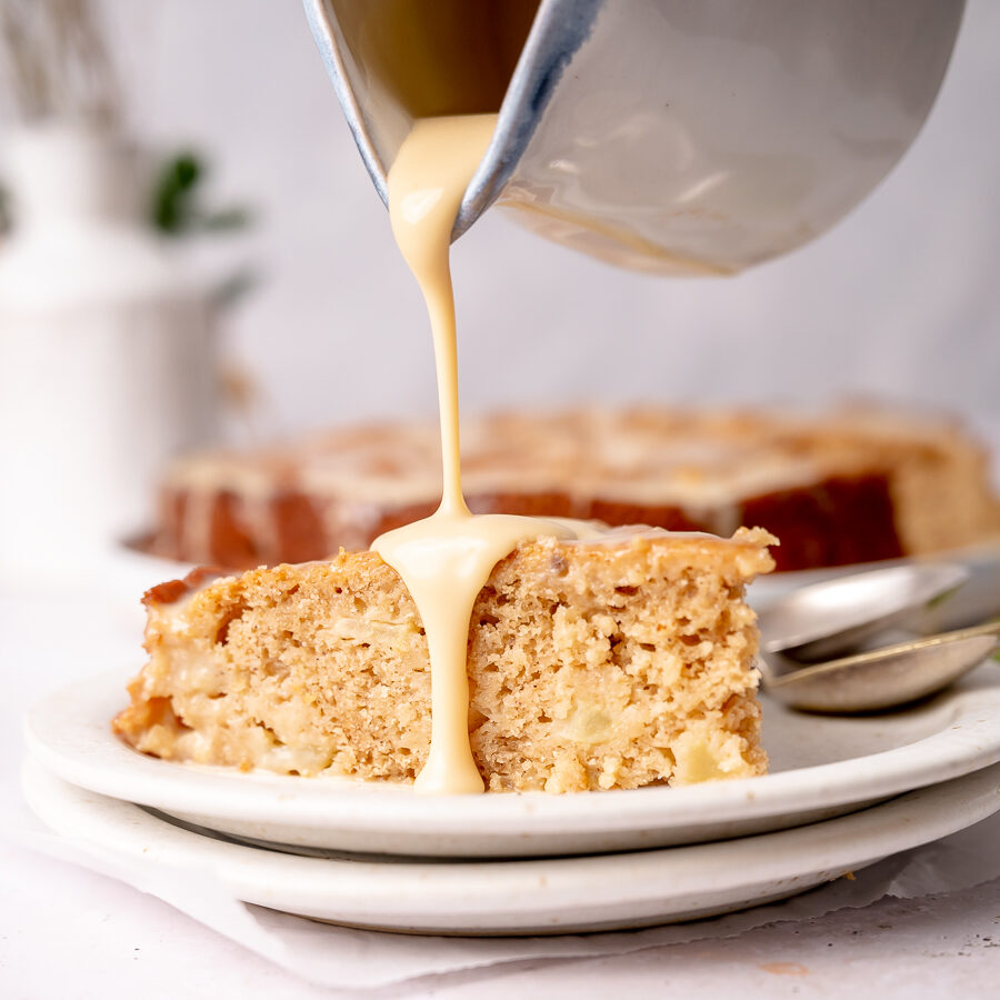 Creamy vanilla sauce slowly being poured over a slice of buttermilk apple cake.