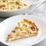 Slice of ham and cheese quiche with parsley sprinkled throughout made in a shallow tart pan as seen in the background.