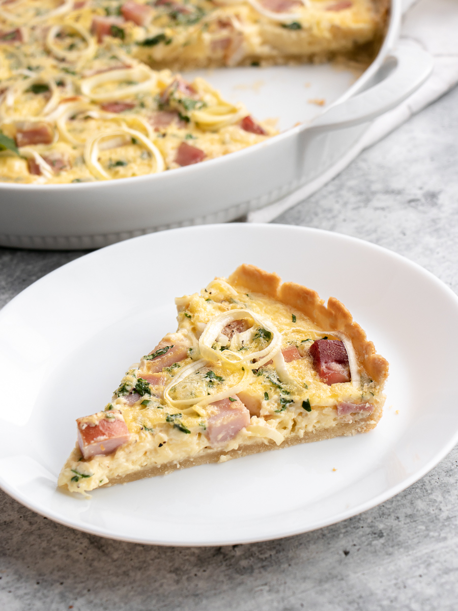 Slice of ham and cheese quiche with parsley sprinkled throughout made in a shallow tart pan as seen in the background.