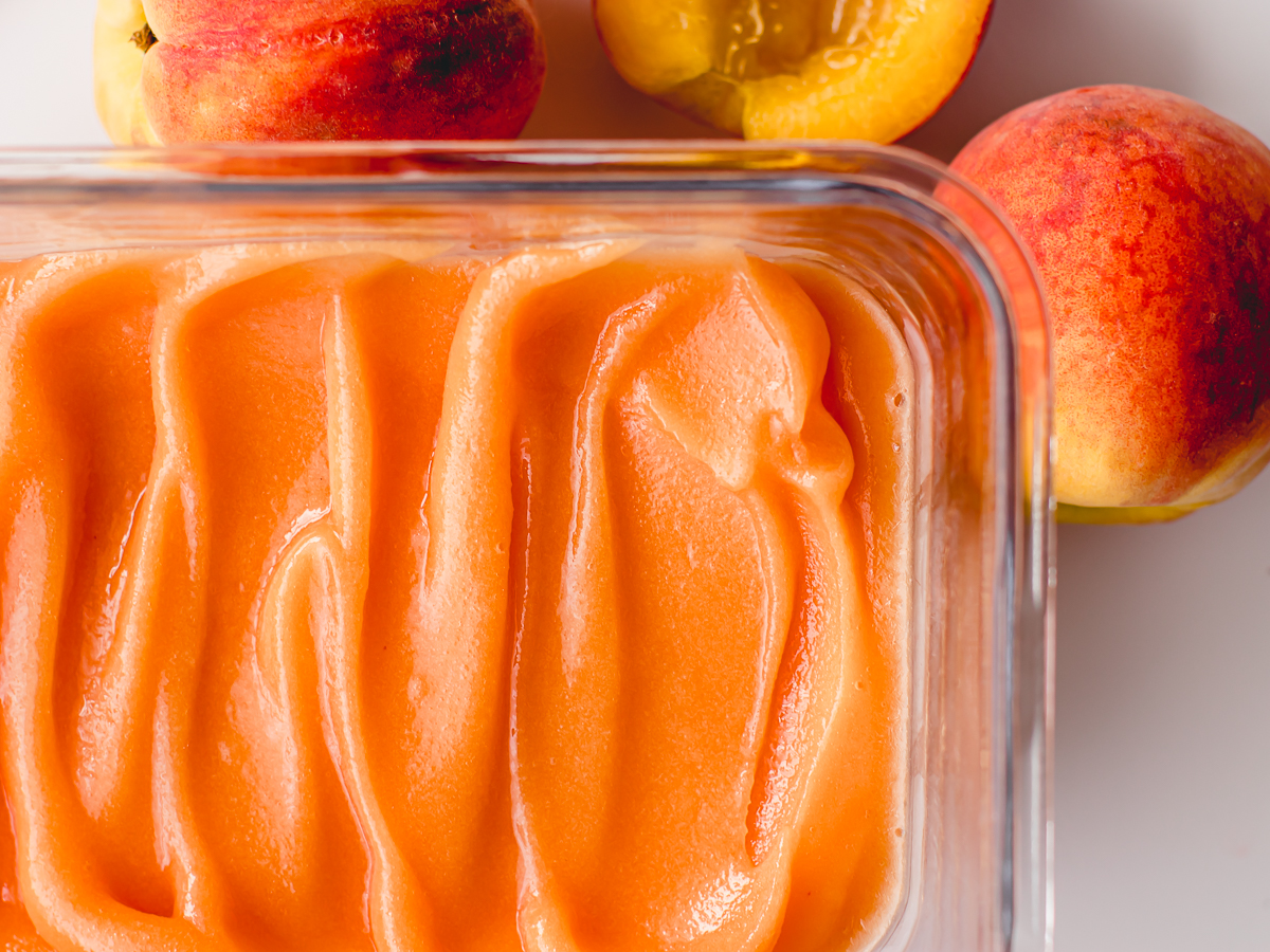 Peach sorbet in a glass container surrounded by fresh peaches.